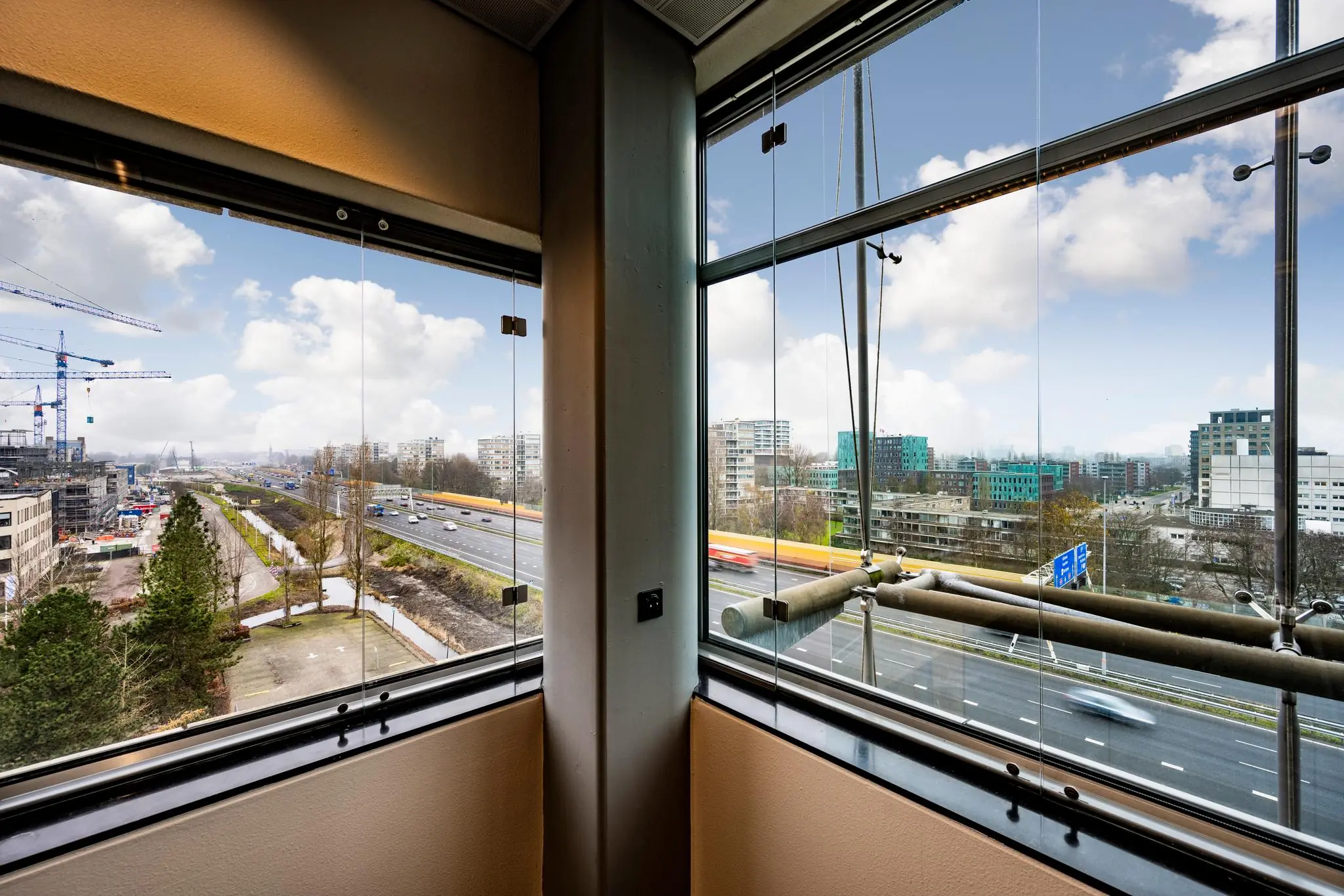 Uitzicht vanuit een hoekraam op de Burgemeester Rijnderslaan met snelweg, hoogbouw en bouwkranen in Amstelveen.